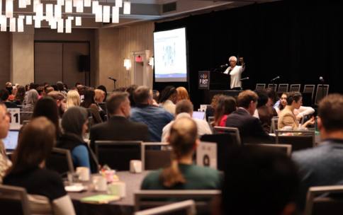 Marie Bernard, M.D., speaks enthusiastically with her hand raised to an audience at the FIRST program conference. She stands next to a projected slide and behind of a podium with the FIRST logo. The room is full and the audience has their backs to the camera, facing Bernard. 