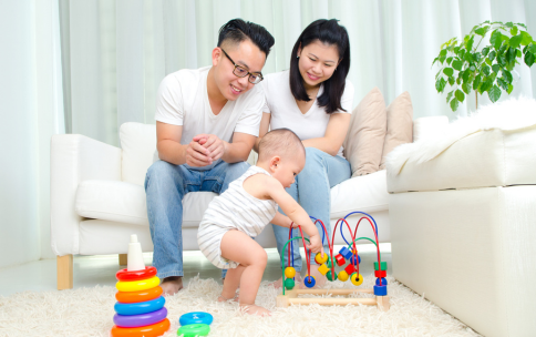 Two parents sit on a sofa watching their infant child playing with toys in front of them