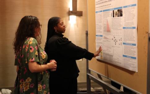 A woman gestures her hand to a poster to show another woman, with her back to the camera, data on the scientific poster.